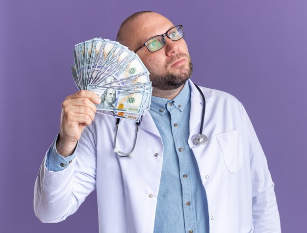 Impressed middle-aged male doctor wearing medical robe and stethoscope with glasses holding money looking at side