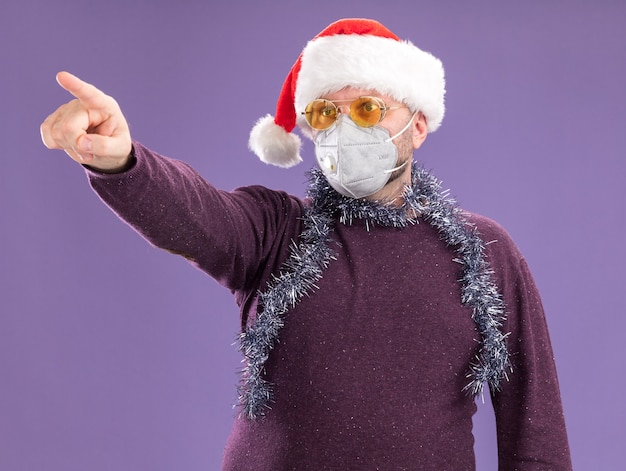 Impressed middle-aged man wearing santa hat and protective mask with tinsel garland around neck with glasses looking and pointing at side isolated on purple wall