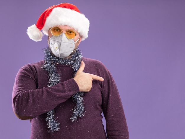 Impressed middle-aged man wearing santa hat and protective mask with tinsel garland around neck with glasses  pointing at side isolated on purple wall with copy space