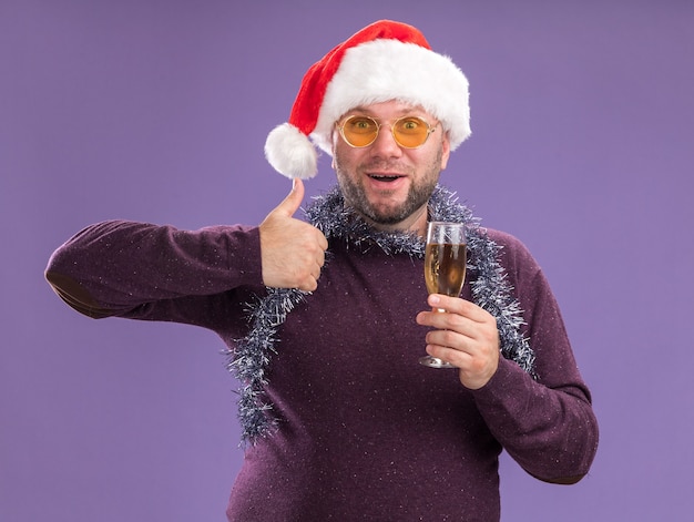 Free photo impressed middle-aged man wearing santa hat and tinsel garland around neck with glasses holding glass of champagne  showing thumb up isolated on purple wall