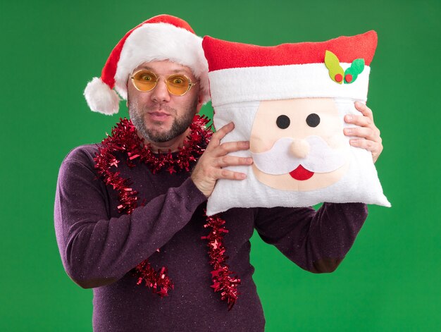 Impressed middle-aged man wearing santa hat and tinsel garland around neck with glasses holding santa claus pillow  isolated on green wall