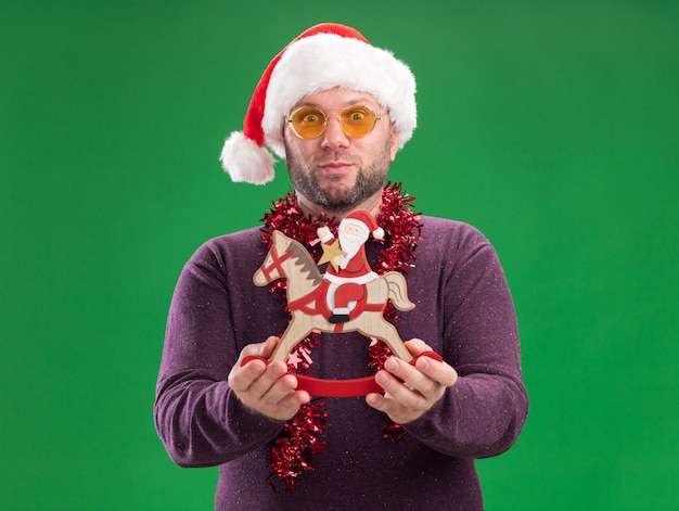 Free Photo impressed middle-aged man wearing santa hat and tinsel garland around neck with glasses stretching out santa on rocking horse figurine   isolated on green wall