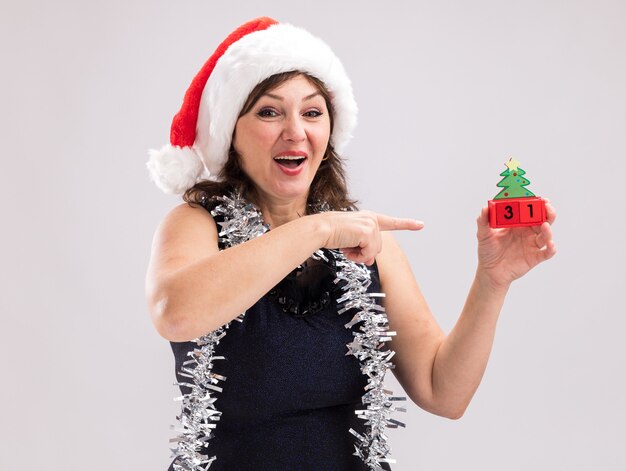 Impressed middle-aged woman wearing santa hat and tinsel garland around neck holding and pointing at christmas tree toy with date looking at camera isolated on white background