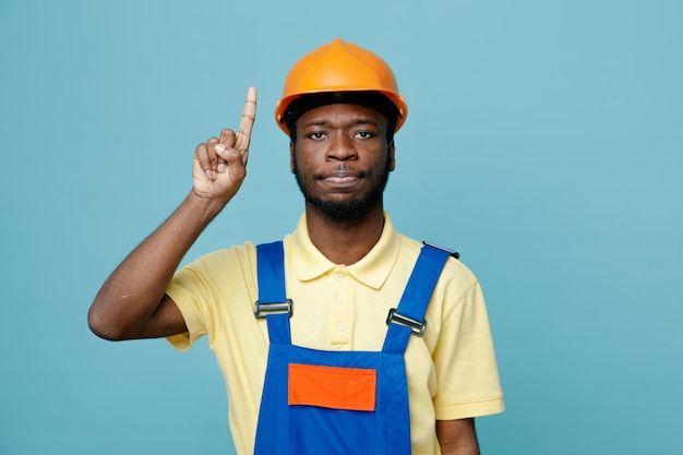 impressed points at up young african american builder in uniform isolated on blue background