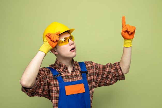 Free photo impressed points at up young male builder wearing uniform and gloves with glasses