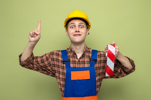 Free photo impressed points at up young male builder wearing uniform holding duct tape