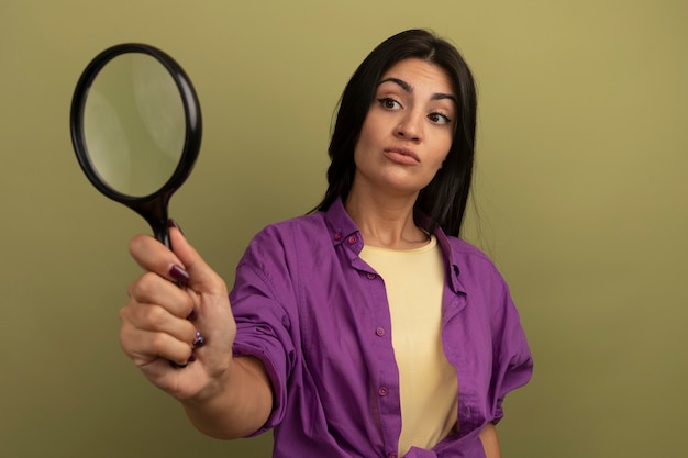 Impressed pretty brunette woman holds and looks at magnifying glass isolated on olive green wall