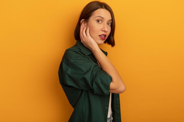 Impressed pretty caucasian woman stands sideways puts hand on face on orange