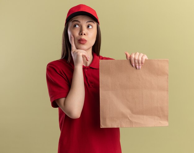 Impressed pretty delivery woman in uniform puts finger on face and holds paper package looking at side on olive green