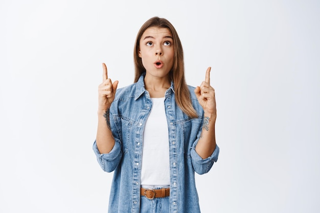 Impressed pretty female model pointing fingers up and raise head at top advertisement checking out something fascinated standing against white background