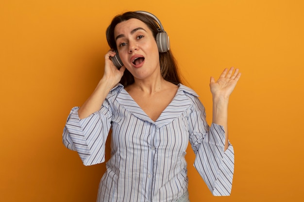 Impressed pretty woman on headphones stands with raised hand isolated on orange wall