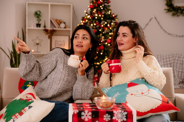 impressed pretty young girls hold cups and look at side sitting on armchairs and enjoying christmas time at home