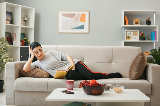 Free Photo impressed putting hand on head young girl reading book lying on sofa behind coffee table in living room