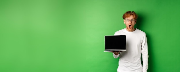 Free photo impressed redhead guy in glasses drop jaw after seeing online promo showing laptop screen and starin