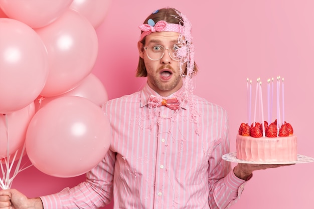 Impressed shocked young man in festive clothes stares bugged eyes keeps mouth opened receives unexpected congratulation from friend smeared with cream holds birthday cake and inflated balloons