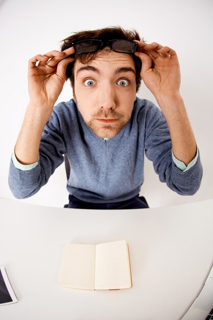 Impressed, and startled young surprised man taking-off glasses, popped eyes while sit at office desk