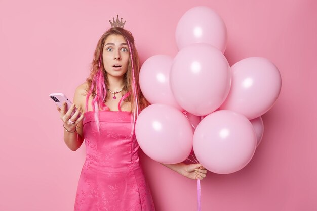 Impressed stunned female model has image of princess wears little crown and dress holds mobile phone gets unexpected message from formal boyfriend comes on party with bunch of inflated balloons