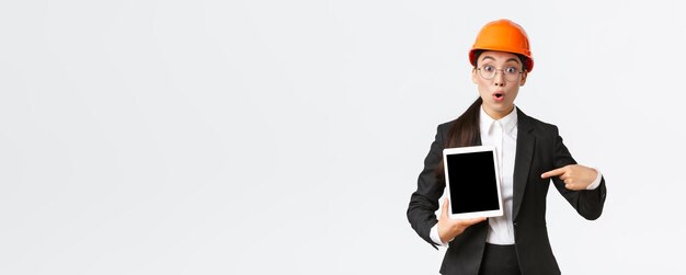 Impressed and surprised asian female engineer technician at industrial area wear safety helmet and business suit pointing finger at profit chart diagram at digital tablet screen look amazed