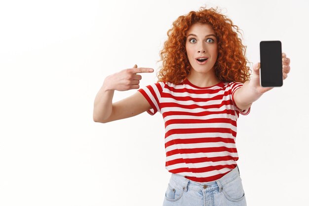 Impressed surprised redhead fascinated young woman curlyhaired ginger girl pointing smartphone display astonished awesome new app recommend cool application standing white background