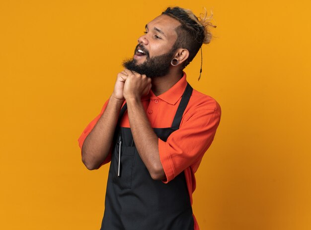 Impressed young afro-american barber wearing uniform keeping hands under chin looking at side