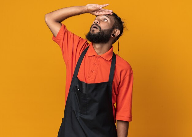 Impressed young afro-american barber wearing uniform touching forehead looking up isolated on orange wall with copy space