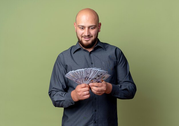 Impressed young bald call center man holding and looking at money isolated on olive green  with copy space