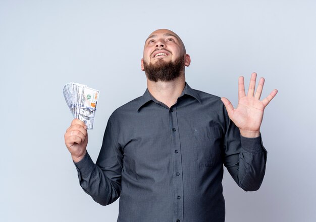 Impressed young bald call center man holding money showing empty hand and looking up isolated on 