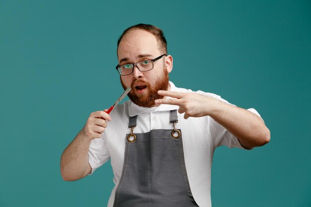 Impressed young barber wearing uniform and glasses showing straight razor looking at camera showing peace sign isolated on blue background