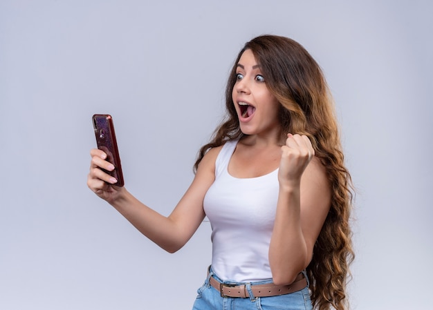 Impressed young beautiful girl holding mobile phone looking at it with raised fist 