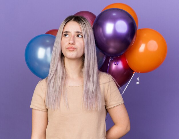 Impressed young beautiful girl standing in front balloons isolated on blue wall