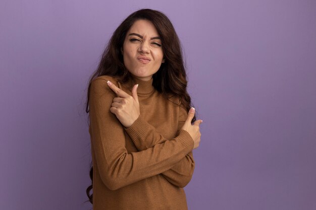 Impressed young beautiful girl wearing brown turtleneck sweater points at differents sides isolated on purple wall with copy space