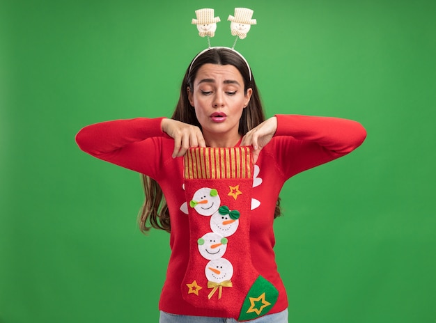 Free photo impressed young beautiful girl wearing christmas sweater with christmas hair hoop holding and looking into christmas socks isolated on green background