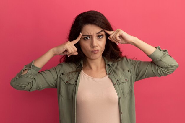 Impressed young beautiful girl wearing olive green t-shirt putting fingers on temple isolated on pink wall