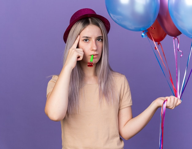 Impressed young beautiful girl wearing party hat holding balloons blowing party whistle putting finger on temple isolated on blue wall