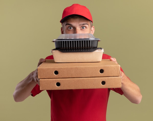 Impressed young blonde delivery boy holds food packages on pizza boxes isolated on olive green wall with copy space