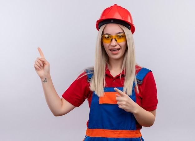 Impressed young blonde engineer builder girl in uniform in dental braces wearing safety glasses pointing at left side on isolated white space