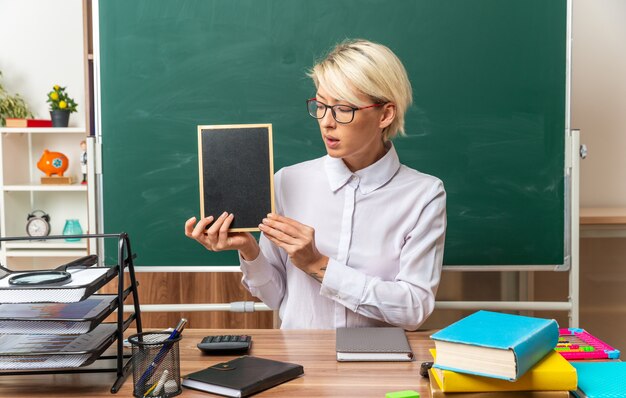 impressed young blonde female teacher wearing glasses sitting at desk with school supplies in classroom showing mini blackboard looking at it