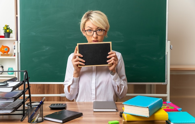 Free photo impressed young blonde female teacher wearing glasses sitting at desk with school tools in classroom showing mini blackboard looking at camera