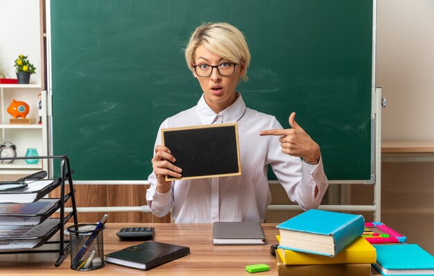 Impressed young blonde female teacher wearing glasses sitting at desk with school tools in classroom showing and pointing at mini blackboard looking at camera