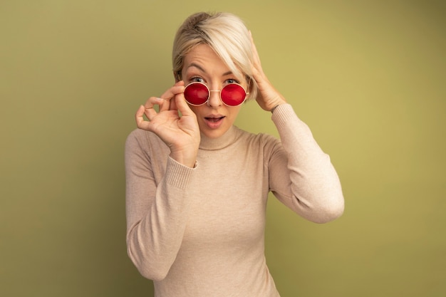 Free Photo impressed young blonde girl wearing and grabbing sunglasses looking  putting hand on head isolated on olive green wall with copy space