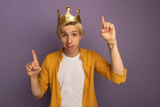 Free Photo impressed young blonde guy wearing yellow t-shirt and crown points at up isolated on purple