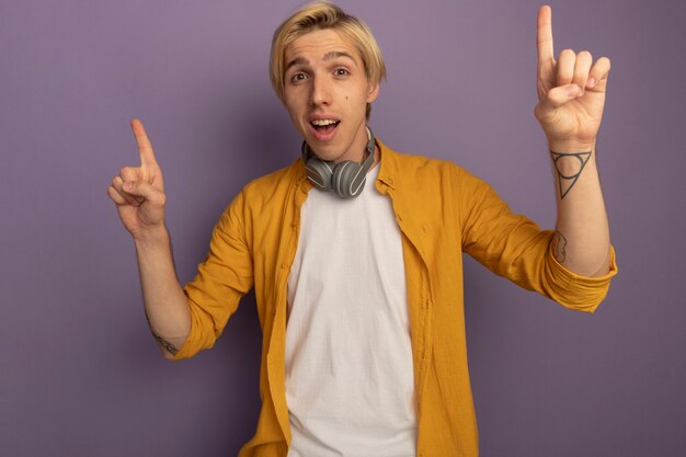 Impressed young blonde guy wearing yellow t-shirt and headphones on neck points at up isolated on purple