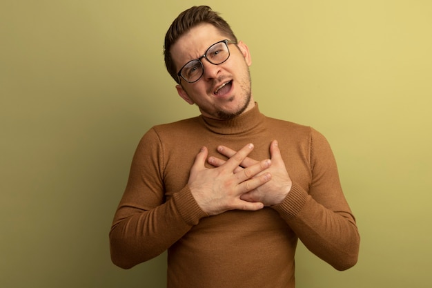 Impressed young blonde handsome man wearing glasses looking  putting hands on chest isolated on olive green wall with copy space