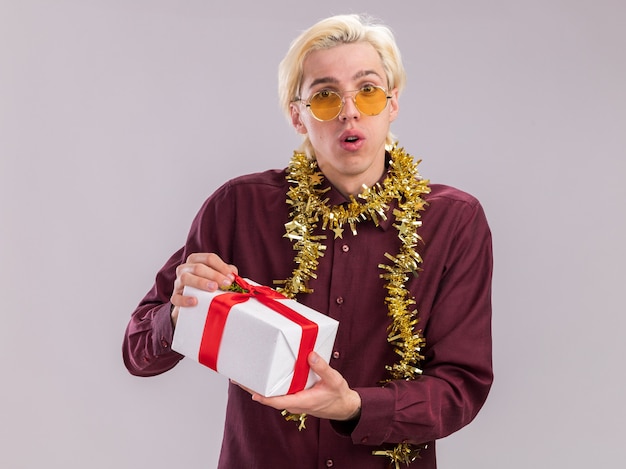 Impressed young blonde man wearing glasses with tinsel garland around neck holding gift package looking at camera isolated on white background