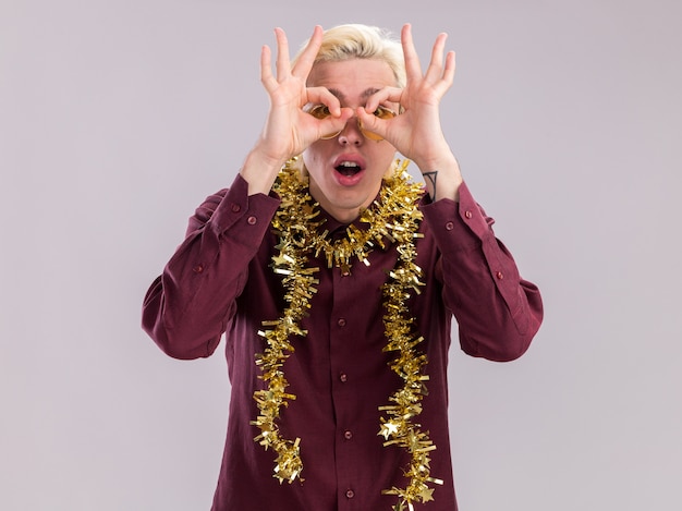 Impressed young blonde man wearing glasses with tinsel garland around neck looking at camera doing look gesture using hands as binoculars isolated on white background