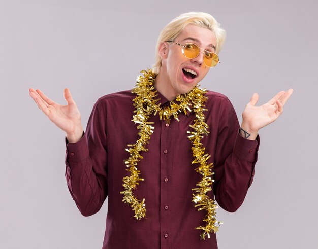 Impressed young blonde man wearing glasses with tinsel garland around neck looking at camera showing empty hands isolated on white background