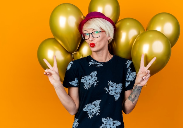 Free photo impressed young blonde party woman wearing party hat and glasses standing in front of balloons doing kiss gesture and peace signs looking at front isolated on orange wall