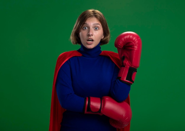 Free photo impressed young blonde superhero woman in red cape wearing box gloves keeping hand in air looking at front isolated on green wall with copy space