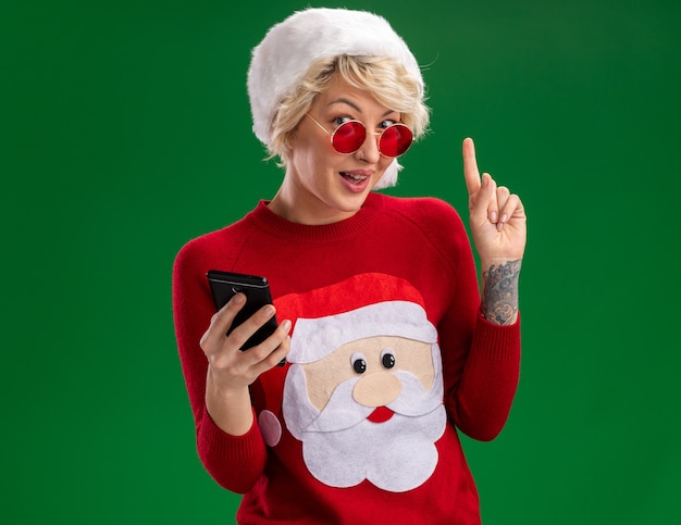 Impressed young blonde woman wearing christmas hat and santa claus christmas sweater with glasses holding mobile phone looking  and pointing up isolated on green wall with copy space