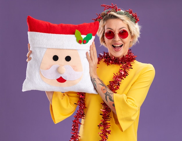 Free photo impressed young blonde woman wearing christmas head wreath and tinsel garland around neck holding santa claus pillow looking  isolated on purple wall